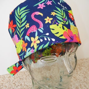 May include: A navy blue scrub cap with a tropical flamingo and floral pattern. The cap features a pink flamingo, colourful flowers, and green leaves. The cap is made of fabric and has a tie at the back.