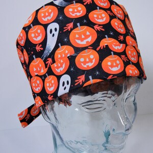 Pode incluir: Um gorro de Halloween com um padrão repetido de abóboras laranja e fantasmas brancos sobre um fundo preto. O tecido também inclui folhas laranja e pequenas estrelas brancas. O gorro é amarrado na parte de trás.