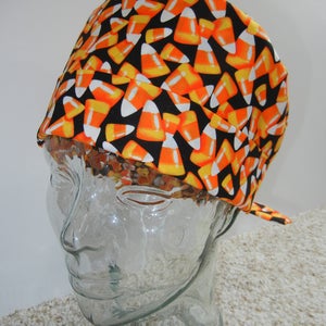 Op de afbeelding: Een zwarte scrub-muts met een snoepmaisprint in oranje, geel en wit. De muts zit op een heldere glazen hoofdform.