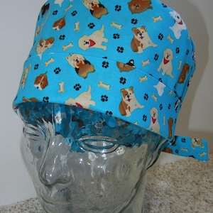 Op de afbeelding: Een blauwe chirurgische scrub-muts met een patroon van honden en potafdrukken. De muts heeft een strik aan de achterkant.