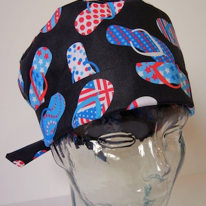 Könnte beinhalten: Eine schwarze OP-Haube mit einem Muster aus roten, weißen und blauen Flip-Flops. Die Flip-Flops haben verschiedene Designs, darunter Punkte, Sterne und Streifen. Die Haube ist am Hinterkopf gebunden.