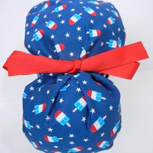 May include: A blue scrub cap featuring a repeating pattern of red, white, and blue popsicle graphics interspersed with small white stars. The cap is gathered at the back and secured with a bright red fabric ribbon tied into a bow.