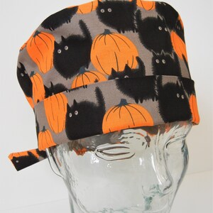 Pode incluir: Um gorro de cirurgia cinza com uma estampa de gato preto e abóbora laranja.