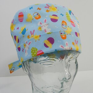 Könnte beinhalten: Eine hellblaue OP-Mütze mit Ostern-Motiv. Die Mütze ist mit einem Muster aus bunten Ostereiern, Hasen, Küken und Schmetterlingen bedruckt.