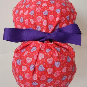 May include: Red fabric scrub hat with a purple ribbon tied around it. The fabric is covered in a pattern of pink and purple candy hearts with various messages, including "XOXO", "HUGS", "LOVE", "SMILE", and "BE MINE".