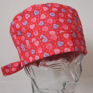 May include: Red and purple heart-shaped patterned scrub hat with a tie. The hearts have various messages on them, including "XOXO", "HUGS", "SMILE", "LOVE", and "CUTE".