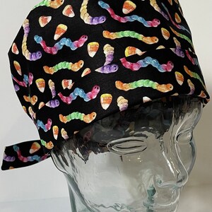 Può includere: Un cappello chirurgico nero con un motivo ripetuto di colorati vermi dei cartoni animati e caramelle a forma di pannocchia. Il cappello è legato sul retro ed è esposto su una testa di manichino di vetro.