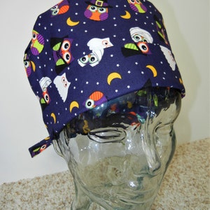 Può includere: Un cappello da scrub viola con una stampa di Halloween di gufi e fantasmi. Il cappello ha un laccio sul retro.
