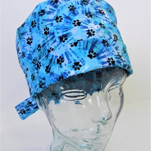 Op de afbeelding: Een blauwe en witte tie-dye scrub-muts met een pootprintpatroon. De muts heeft een strik aan de achterkant.