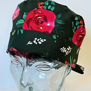 Può includere: Un cappello da scrub verde scuro con una stampa floreale con rose rosa e rosse, foglie verdi e dettagli bianchi.