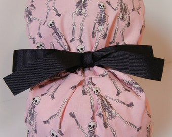 Könnte beinhalten: Eine rosa Stoff-OP-Mütze mit einer schwarzen Schleife. Die Mütze ist mit einem sich wiederholenden Muster aus tanzenden schwarzen Skeletten verziert.