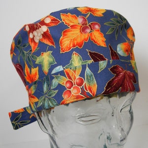 Pode incluir: Um gorro cirúrgico azul com uma estampa colorida de folhas de outono e bagas. O gorro tem uma gravata que pode ser amarrada na parte de trás do pescoço.
