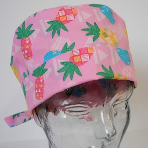 May include: Pink surgical scrub hat with a pineapple print. The hat has a tie on the side.