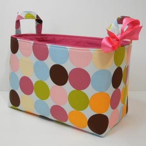 May include: Rectangular fabric storage bin with a colorful polka dot pattern in pink, blue, green, yellow, orange, and brown. The bin has two handles and a pink interior. A pink bow is tied on one handle.