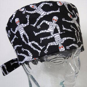 Può includere: Cappello da lavoro nero con un motivo di mummia bianco e arancione. Il cappello ha una cravatta sul lato.