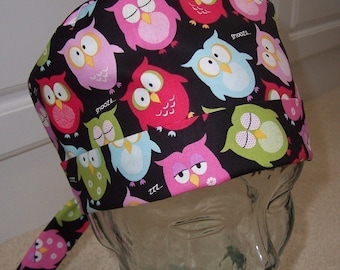 May include: A black scrub hat with a colorful owl print. The owls are pink, blue, green, and yellow with black eyes. The hat has a tie that is pink with white flowers.