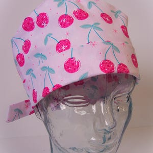 May include: A pink surgical scrub hat with a pattern of pink cherries with a disco ball design on a white background.