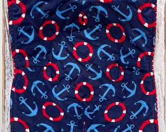 May include: A navy blue quilted bowl cozy with a nautical theme. The fabric features a pattern of light blue anchors and red and white life preservers. The edges are trimmed with red binding. The cozy is designed to hold a bowl of hot food.