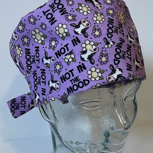 May include: Purple scrub hat with a white daisy and cow print pattern. The text "Not in the mood" is repeated throughout the hat.