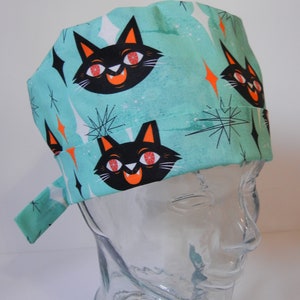Pode incluir: Um gorro de cirurgia azul turquesa com uma estampa de gato preto. O gato tem olhos laranja e um sorriso branco. O gorro tem uma amarração lateral.