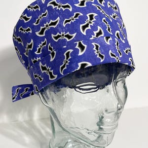 May include: A scrub cap featuring a vibrant purple background with a repeating pattern of black and white bats. The cap has a folded front band and a tie-back closure at the rear, providing a secure fit for the wearer while maintaining a consistent look.