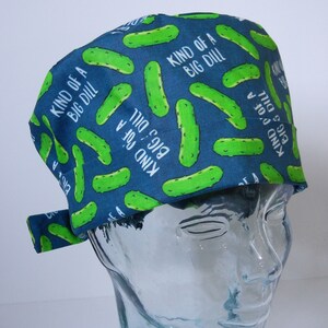 May include: A blue scrub cap featuring a pattern of green pickle illustrations and the white text "KIND OF A BIG DILL". The cap is tied at the back and is displayed on a clear mannequin head.