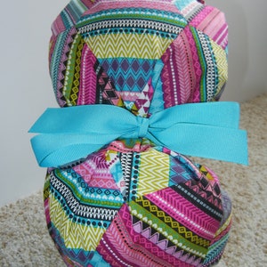 May include: A colorful patterned fabric scrub hat with a turquoise bow. The hat has a geometric design with pink, green, blue, and black stripes and patterns.