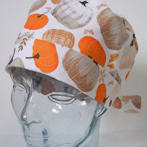 Pode incluir: Um gorro de cirurgia branco com uma estampa de outono de abóboras laranja e folhas marrons. O gorro tem um laço na parte de trás.