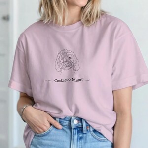 Puede incluir: Camiseta rosa claro con un dibujo lineal de la cara de un perro Cockapoo y el texto "Cockapoo Mum" en negro. La camiseta es de material suave y se combina con unos vaqueros azules.