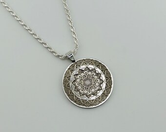 May include: A silver pendant necklace with a detailed mandala design. The pendant has a circular shape with intricate patterns and textures. The chain is a delicate rope style. The necklace is displayed against a light background.