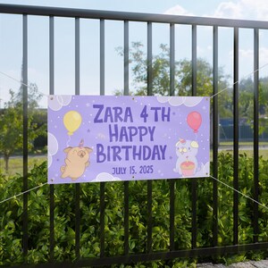 Może przedstawiać: Fioletowy baner urodzinowy z napisem "ZARA 4TH HAPPY BIRTHDAY JULY 15, 2025". Baner zawiera ilustracje kreskówkowe psów z rogami jednorożca, balonami i chmurami. Baner wisi na czarnym metalowym płocie.