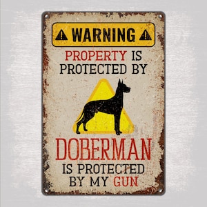 May include: A yellow and black warning sign with the text "WARNING PROPERTY IS PROTECTED BY DOBERMAN IS PROTECTED BY MY GUN". The sign features a silhouette of a Doberman Pinscher dog inside a yellow triangle.