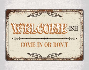 May include: A rusty metal sign with a white background and brown text. The sign says "Welcome-ish Come in or Don't" with a decorative arrow and feather design.