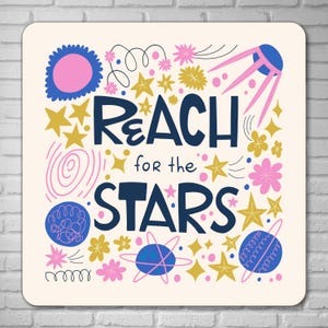 May include: A square, cream-coloured print featuring the navy blue text "REACH for the STARS". The design includes gold stars, pink and yellow flowers, and a blue and pink satellite illustration. The overall theme is inspirational.