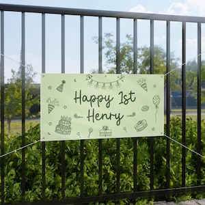 Może przedstawiać: Jasnozielony baner z napisem "Happy 1st Henry" w stylu ręcznym, ozdobiony ilustracjami o tematyce urodzinowej. Baner jest zawieszony między dwoma punktami na czarnym metalowym płocie, z zielenią w tle.