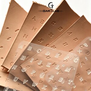 May include: A set of tan leather sheets featuring embossed alphabet letters and numbers, accompanied by a clear plastic stencil template displaying the full alphabet and numbers. The logo GARTLAB is printed in black at the top center.