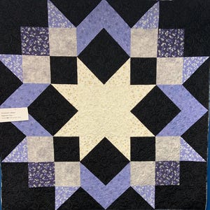 May include: A handmade quilt with a starburst design. The quilt features a central cream-colored star, surrounded by black, lavender, and gray geometric shapes. Small floral patterns accent the design. The quilt is likely made of cotton and has a quilted texture.