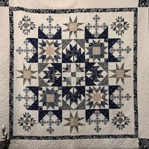 Op de afbeelding: Een handgemaakte quilt met een ster- en geometrisch patroon in tinten wit, marineblauw en bruin. De quilt heeft een gedetailleerd ontwerp met verschillende stervormen en bloemaccenten, perfect voor een bed of wandkleed.