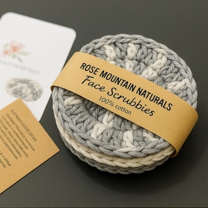 May include: Three gray and white crocheted face scrubbies with a brown paper label that reads "Rose Mountain Naturals Face Scrubbies 100% cotton".