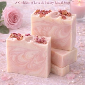Puede incluir: Barras apiladas de jabón artesanal con un diseño en espiral rosa y blanco, adornadas con pétalos de rosa secos. El jabón está etiquetado como "Aphrodite's Embrace" y "Rose Mountain Naturals". Una rosa y una vela encendida en el fondo.