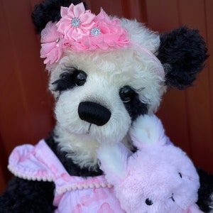 May include: A black and white panda bear wearing a pink flower headband and a pink dress with a floral pattern. The bear is holding a pink bunny rabbit plush toy.