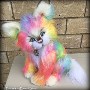 May include: A colorful, plush cat-like creature with rainbow-colored fur, blue eyes, and a pink nose. The handmade stuffed animal has white paws, ears, and tail. The text "Fantasia' by Emma's Bears" is at the bottom.
