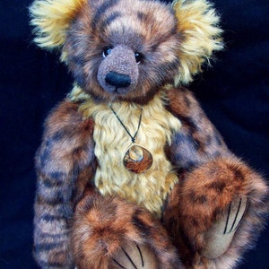 May include: A brown and tan teddy bear with a striped pattern and a brown nose. The bear is wearing a necklace with a brown pendant. The bear is sitting on a person's hand.