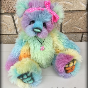 Puede incluir: Un oso de peluche colorido y esponjoso con una diadema rosa de lunares blancos y un collar con un dije rosa. El oso tiene un patrón de pelaje de colores arcoíris y está sentado sobre una superficie blanca. 'Ivy' by Emma's Bears está escrito debajo del oso.