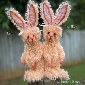 May include: Two handmade plush bunny rabbits, one named 'Lulu' and the other 'Kelbie', are sitting on a wooden fence post. They are both light brown with pink ears and bows. The rabbits are made by 'Emma's Bears'.
