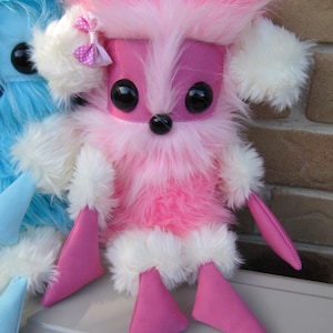 Op de afbeelding: Een roze en wit pluche speelgoed met grote zwarte ogen en roze benen. Het speelgoed heeft een witte strik op zijn hoofd.