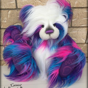 May include: A white teddy bear with blue, pink, and purple fur. The bear has a gray nose and purple eyes. The bear's paws are purple with embroidered paw prints. The bear is sitting on a white surface. 'Gemini' by Emma's Bears.
