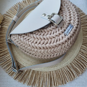 May include: A beige crocheted bag with a white leather flap and a silver buckle. The bag has a long strap and a small label that says "zapletana".