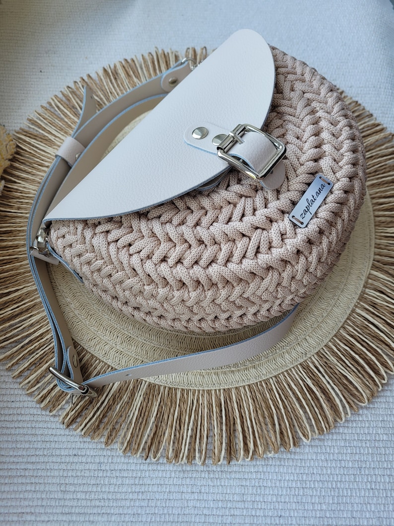 May include: A beige crocheted bag with a white leather flap and a silver buckle. The bag has a long strap and a small label that says "zapletana".