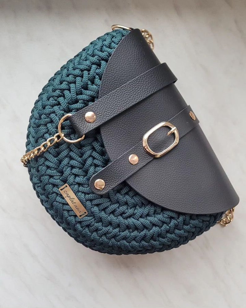 May include: Teal crochet handbag with a black leather flap, gold buckle and rivets. The bag has a gold chain strap and a small gold tag reading "zaplatana".